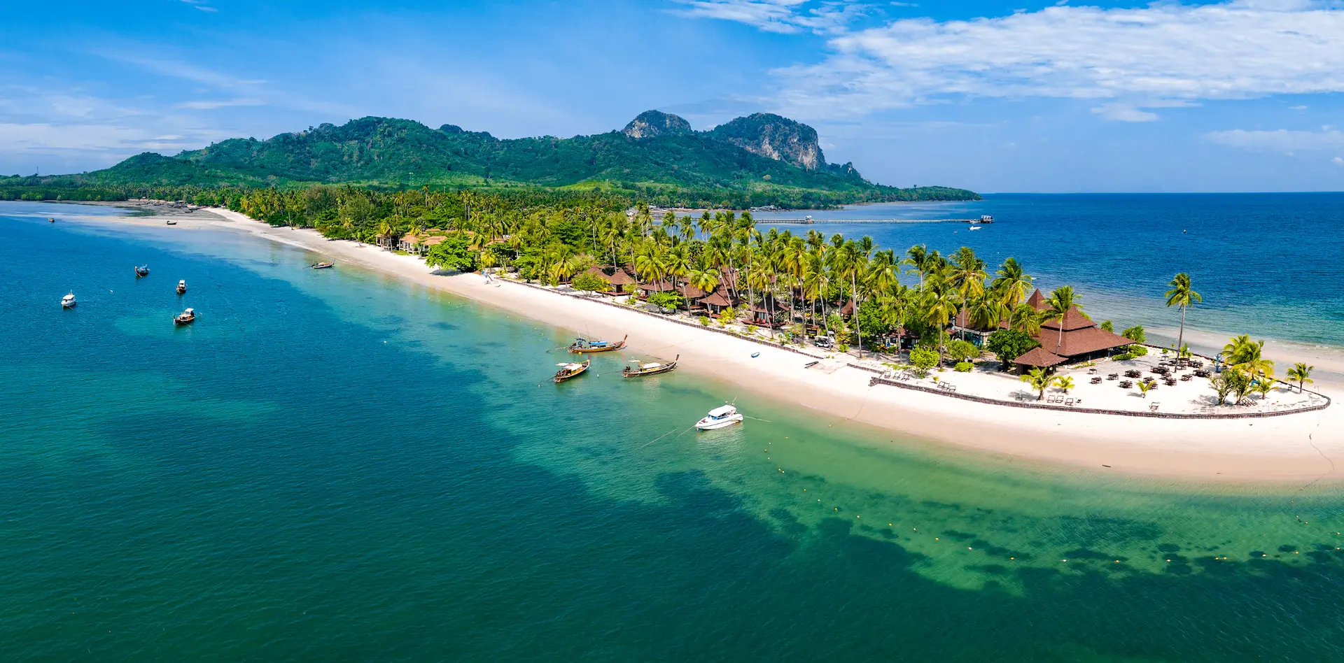 A tropical island with a narrow sandy beach, palm trees, and turquoise water dotted with boats; lush green hills in the background under blue sky.