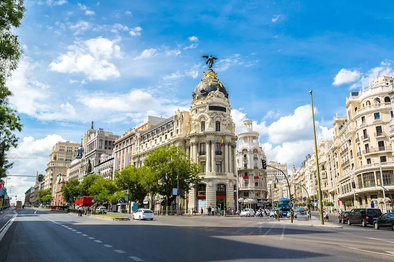 Ornate domed building with a statue on top, at a busy Madrid street corner under a blue sky