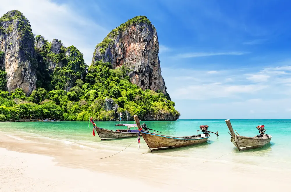 Three wooden long-tail boats tied near a white-sand beach with turquoise water and towering green limestone cliffs in the background.